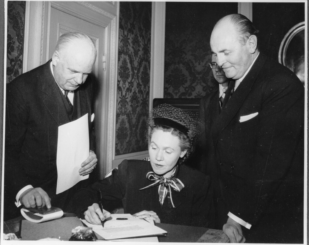Anderson sitting at a table next to two men standing signing the treaty