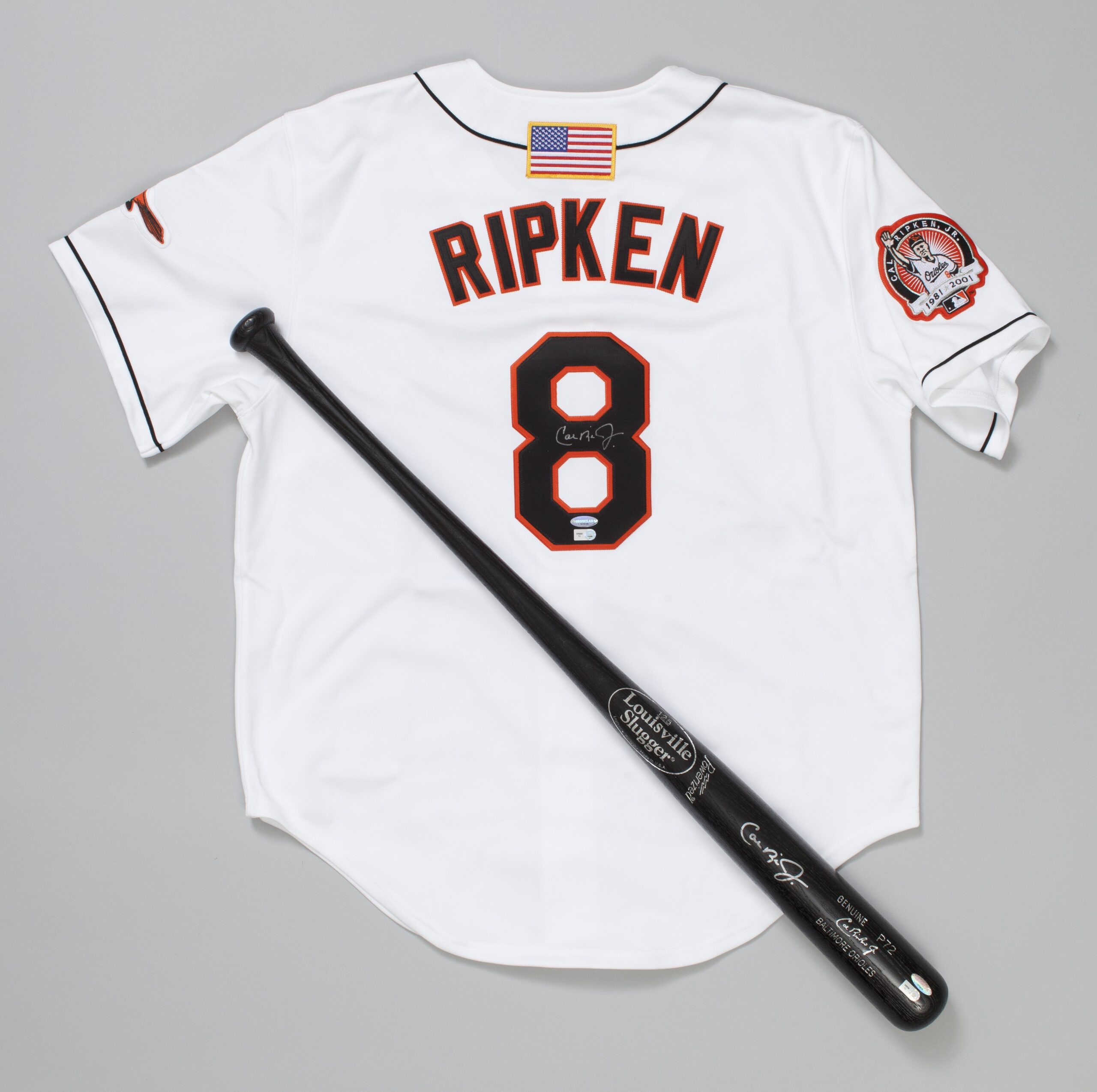 White baseball jersey with text displaying the name "Ripken" and a number 8. Black baseball bat laying on top of jersey. 
