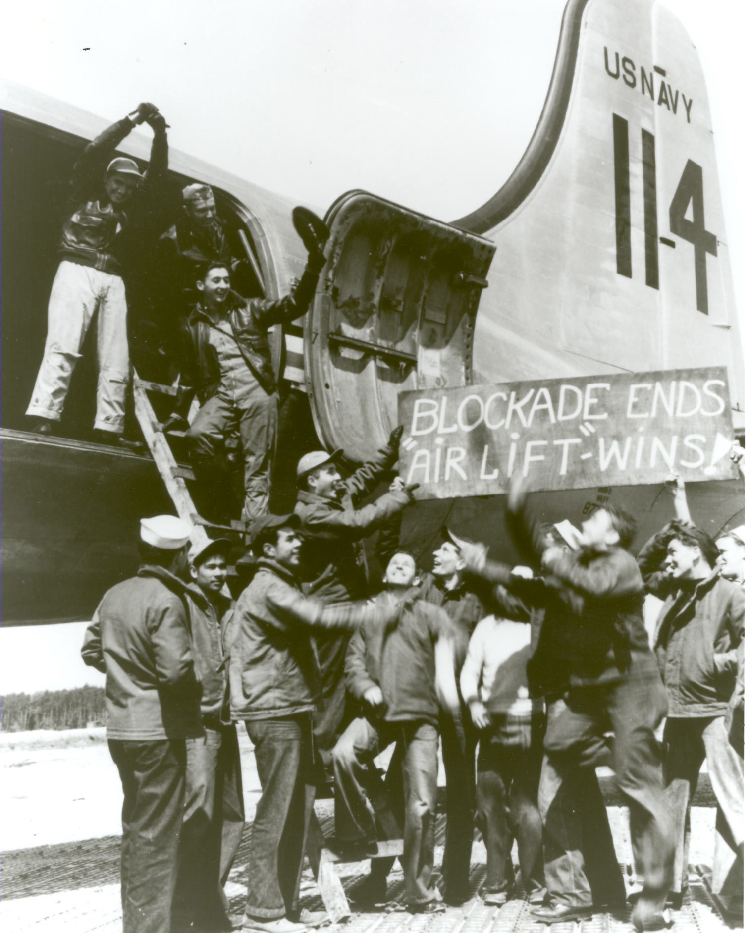 A group of men point to a sign outside of a plane that says "Blockade Ends Air Lift Wins"