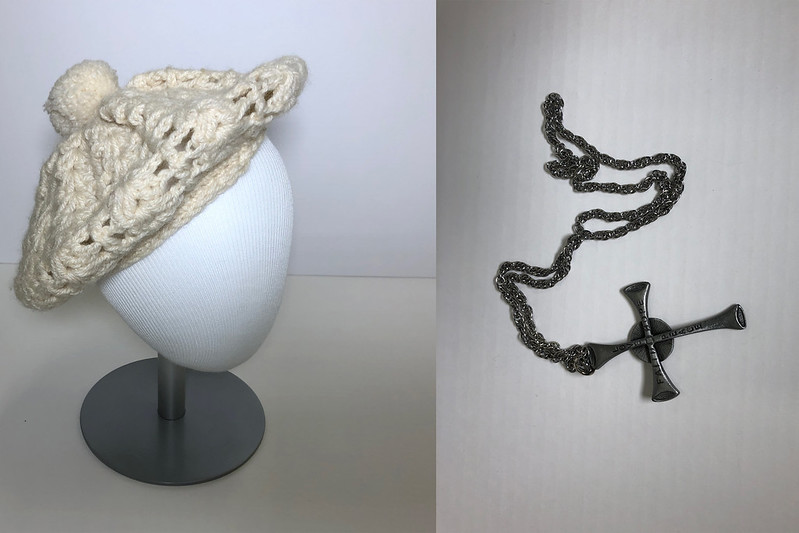 White knitted beret staged on a mannequin head and a dark gray metal cross necklace.