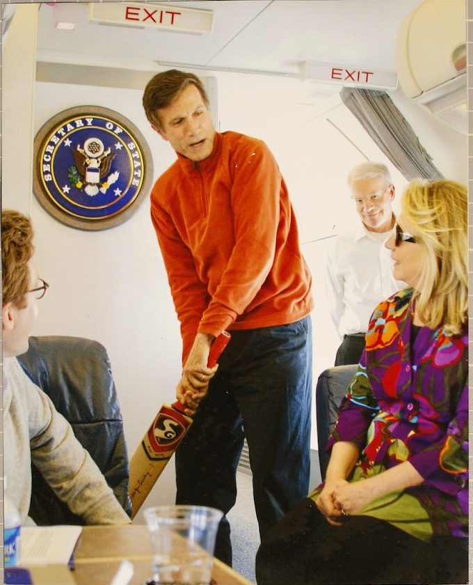 A man in an orange sweater stands in front of a state department seal, holding a cricket bat and talking to a seated man and woman. 