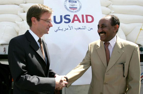 Two men shake hands in front of a large pile of sacks of supplies covered by a "USAID" banner