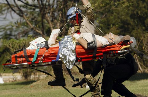 An older injured man lies on a gurney held in the air by a wire from an unseen helicopter above and out of frame. Two workers attend to him.