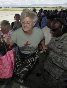 A crowd of people stand in line on an airport tarmac facing the camera. An older woman wearing a green shirt lifts a pink bag ahead of her as she boards the plane.