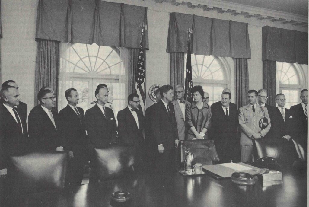 Jean Wikowski stands in a room surrounded by a large group of men in suits.