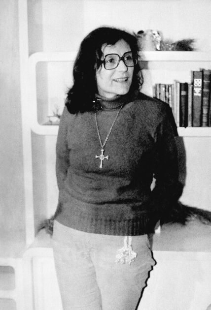 A woman with dark hair stands in front of bookshelf while wearing a cross necklace.