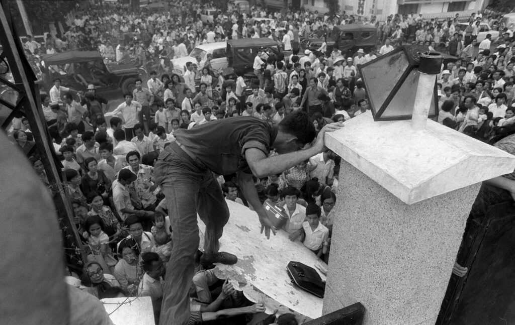 Un gran grupo de personas fuera de una embajada en Vietnam intentando escalar el muro, imagen en blanco y negro.