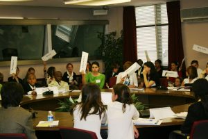 A group of students seated around a table in a classroom, all holding up pieces of cardboard with writing on them