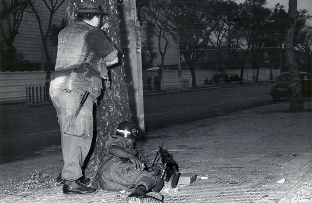 A man standing and a man on the ground in uniform with guns outside the embassy building