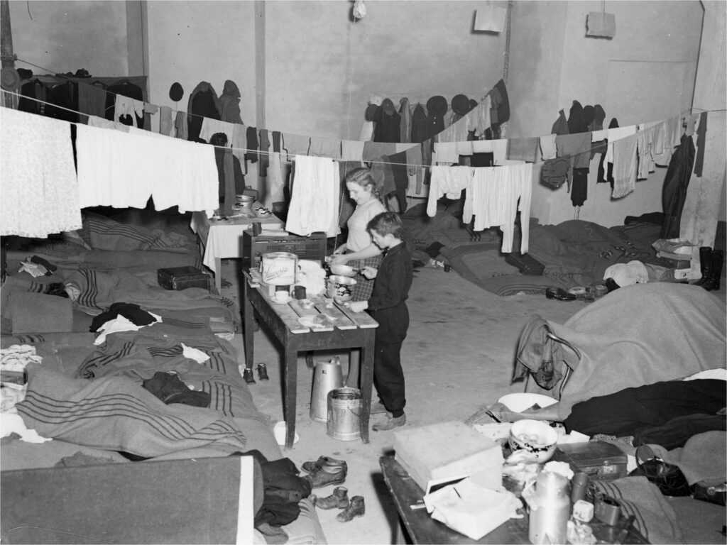 a group of east german refugees in a room surrounded by hanging clothes and sleeping bags