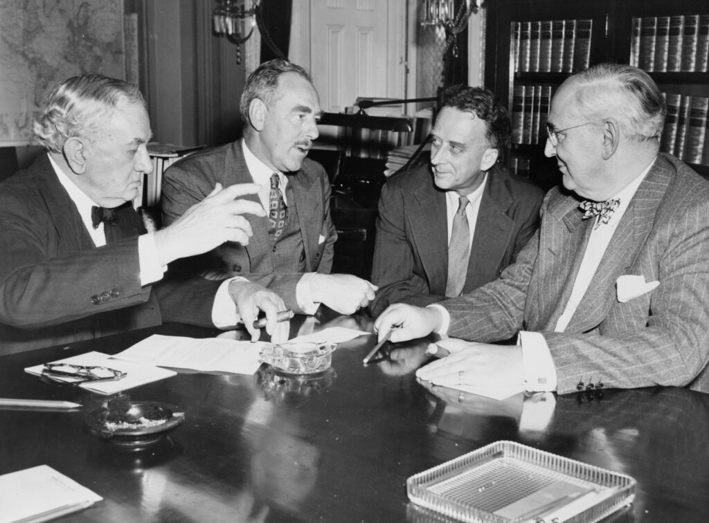 Four men in suits talk around a desk 