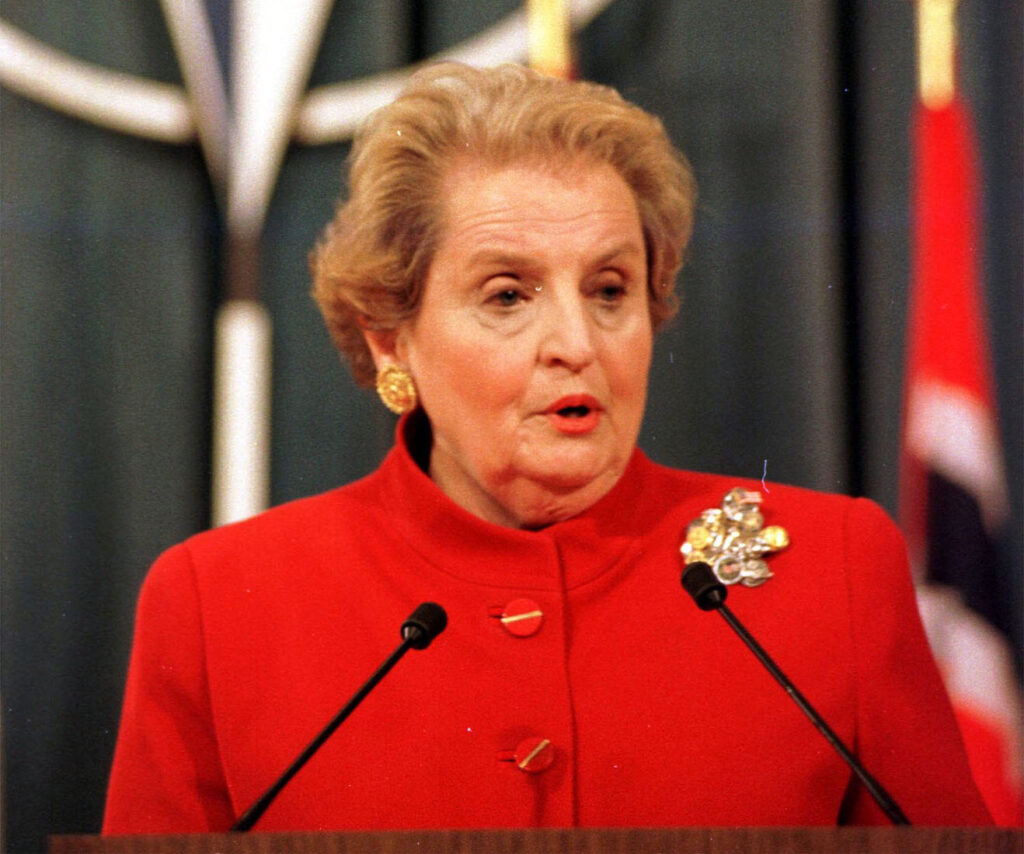 Secretary Albright speaking at a podium in a red jacket