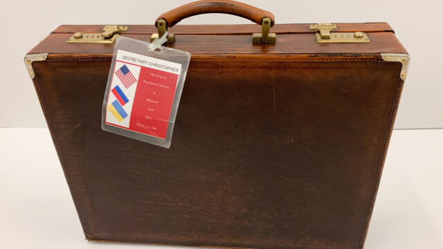 A brown leather briefcase with a tag that says “Secretary Christopher: The Trip of President Clinton to Moscow and Kiev May 8-12, 1995”