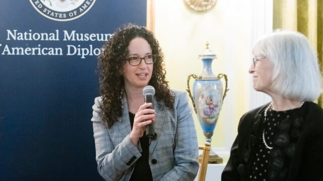 Debora Cahn speaking with Ambassador Beth Jones at an event in front of a National Museum of American Diplomacy banner