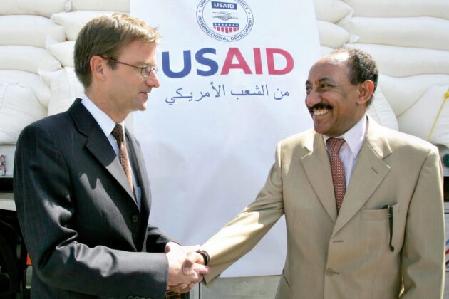 Two men shake hands in front of a large pile of sacks of supplies covered by a “USAID” banner