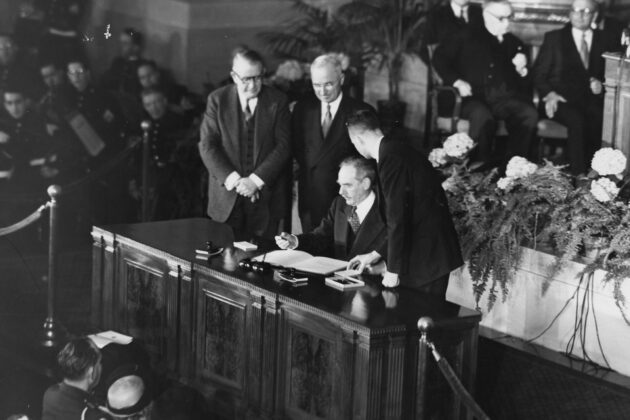 President Truman and Dean Acheson sign the North Atlantic Treaty on a desk in front of an audience