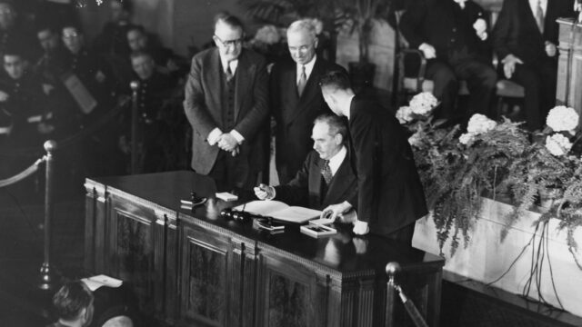 President Truman and Dean Acheson sign the North Atlantic Treaty on a desk in front of an audience