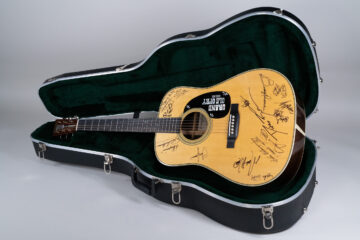 A wooden acoustic guitar covered with autographs in black ink in a black case.