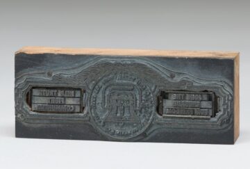 Foto: Druckplatte mit Glocke darauf, links und rechts von der Glocke eine Inschrift