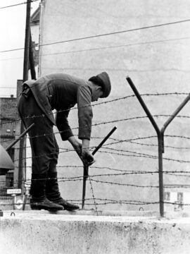 Foto: auf der Mauer stehender ostdeutscher Soldat befestigt Stacheldraht an einer Eisenstange