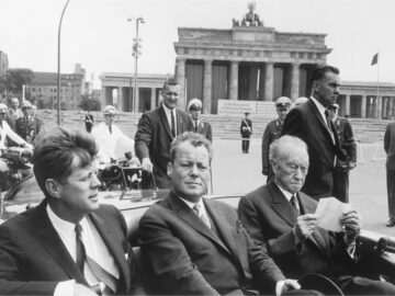 Foto: John F. Kennedy, Willy Brandt und Konrad Adenauer sitzen in einem offenen Wagen, im Hintergrund das Brandenburger Tor