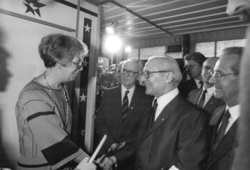 Foto: US-Botschafterin Rozanne Ridgway begrüßt Erich Honecker mit einem Händedruck, hinter ihm stehen Männer