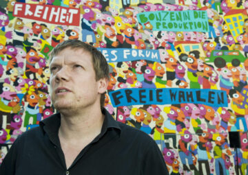 Foto des Künstlers Michael Fischer-Art vor einem Graffito mit den Inschriften „Freiheit“, „Freie Wahlen“, „Bonzen in die Produktion“
