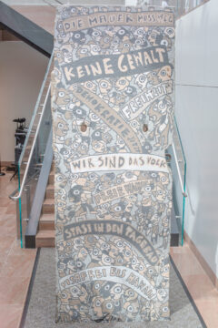 Nahaufnahme eines großen Mauersegments, das in einem Gebäude ausgestellt ist, mit Inschriften wie „Die Mauer muss weg“, „Keine Gewalt“, „Wir sind das Volk“, „Stasi in den Tagebau“, „Visafrei bis Hawaii“ und „Demokratie jetzt“