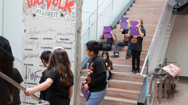 A group of students holding iPads sitting on the stairs of the museum in front of a piece of the Berlin Wall