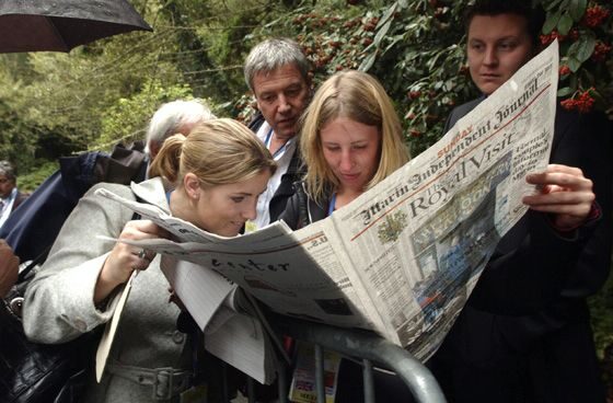 Five adults of varying ages read a newspaper together outdoors.