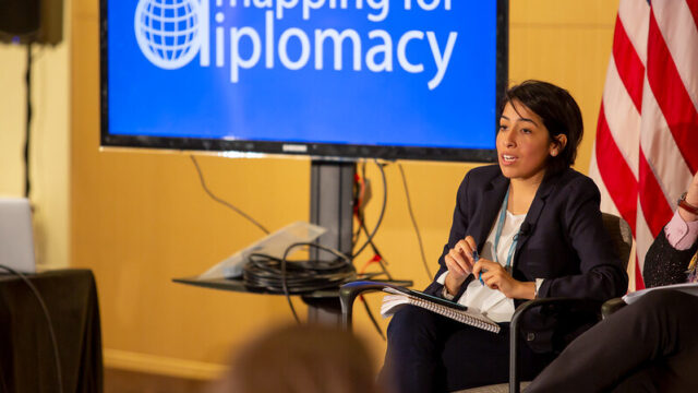 woman sitting in chair addressing audience with screen displaying the words “mapping for diplomacy” behind her.