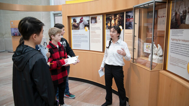 Teacher stands in front of museum exhibit as students listen with clipboards.