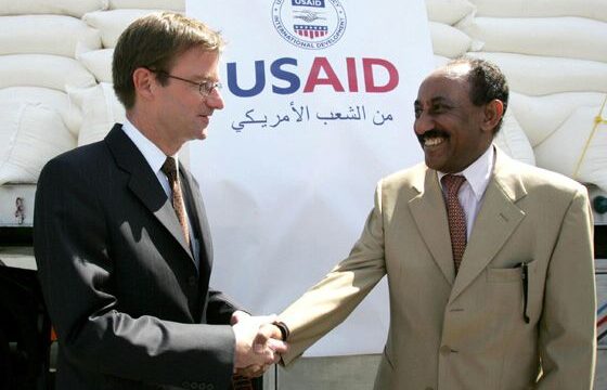 Two men shake hands in front of a large pile of sacks of supplies covered by a “USAID” banner