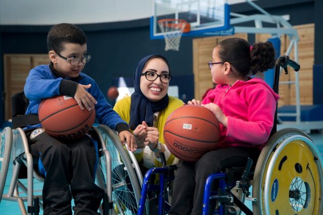 a woman wearing a headscarf and glasses kneeling between two children in wheelchairs holding basketballs on an indoor basketball court