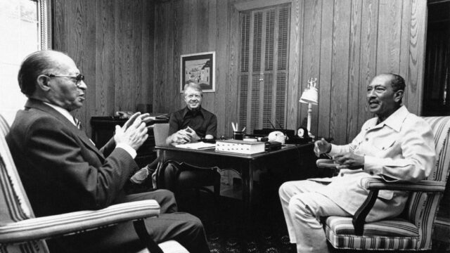 Black and white photo of Begin, Sadat, and Carter sitting in office and talking.