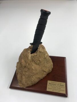 A wooden plaque with a black dagger stabbed into a fake stone on it.
