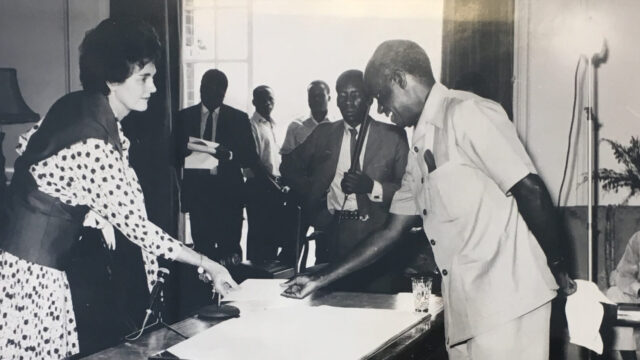 Jean Wilkowski handing papers over a desk to a man standing across from her.