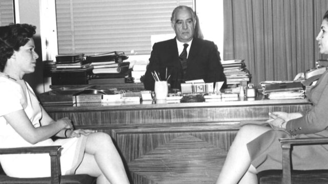 Two women sit across from each other in chairs. Behind them sits a man at a desk.