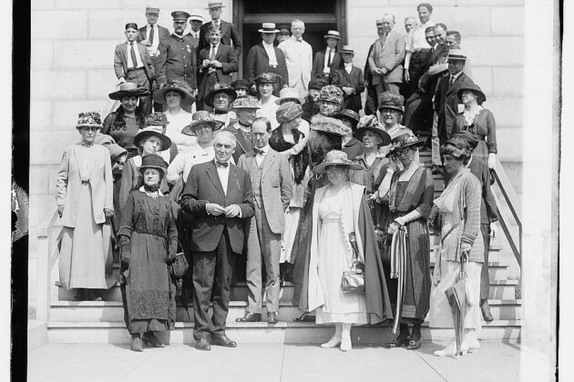A group of women and men stand on the steps in front of a building posing in formal attire for a photograph.