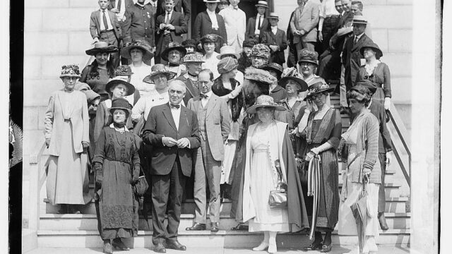 A group of women and men stand on the steps in front of a building posing in formal attire for a photograph.