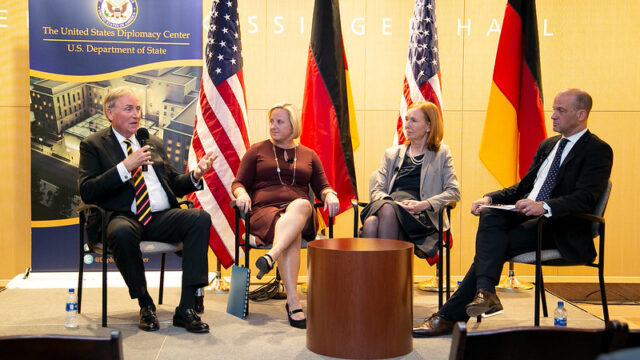 Four panels speaking on stage with American and German flags in the background.