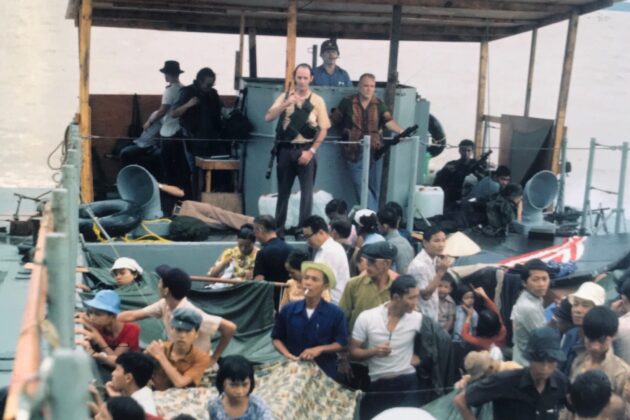 Consul General Francis Terry McNamara (behind the two armed guards) captains a barge of American and Vietnamese refugees out of Cần Thơ.
