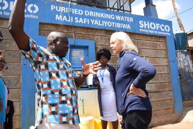 Man speaks with hands to a woman listen with a sign in the background reading “purified drinking water”