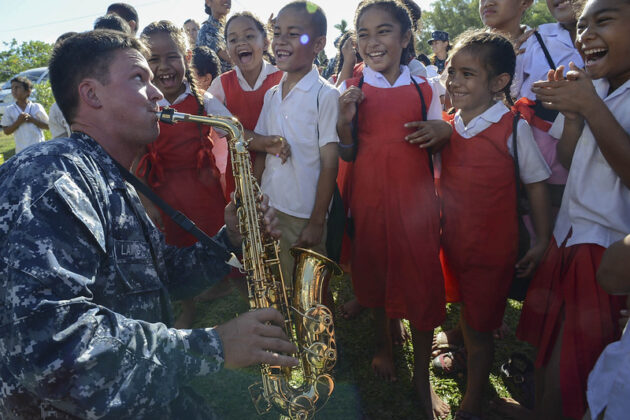 US solider in camouflage uniform plays a saxophone for a group of school children smiling