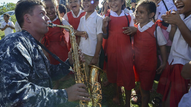 US solider in camouflage uniform plays a saxophone for a group of school children smiling